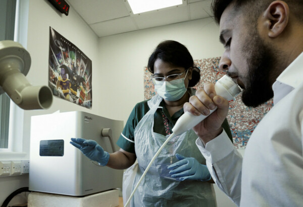 A healthcare professional wearing a dark green uniform, white apron, blue gloves and mask is in a clinic room with a research participant who is wearing a white shirt. The research participant is blowing into a white tube.