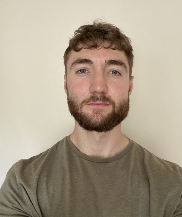 A portrait photograph of a  man with short brown hair, trimmed beard, and blue eyes, wearing an olive-green t-shirt, standing against a plain light-colored background