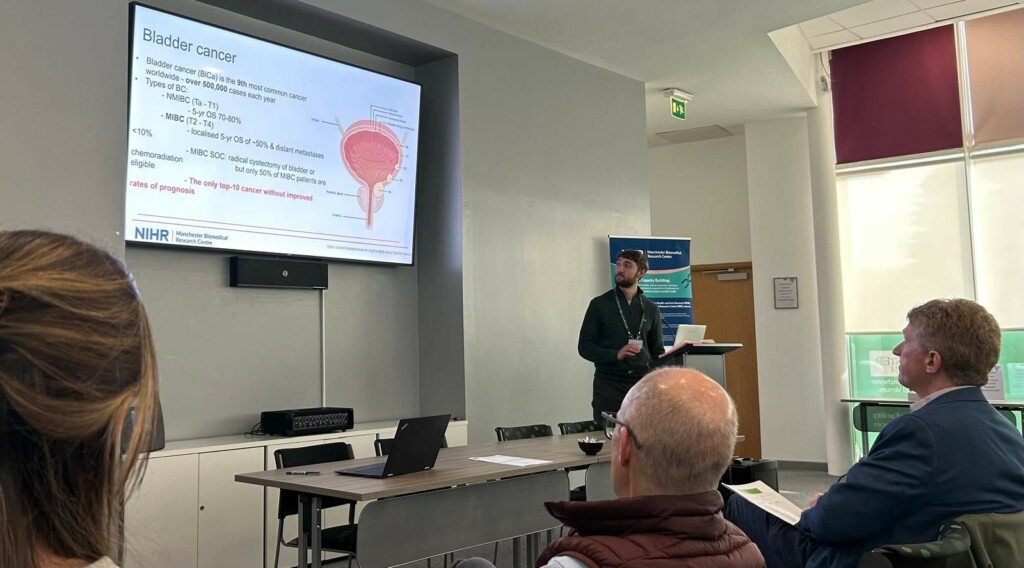 A man giving a presentation with a slide titled 'Bladder cancer' displayed on a large screen. The slide contains text and a diagram of the bladder. The presenter stands near a podium with a laptop, addressing an audience seated at tables.