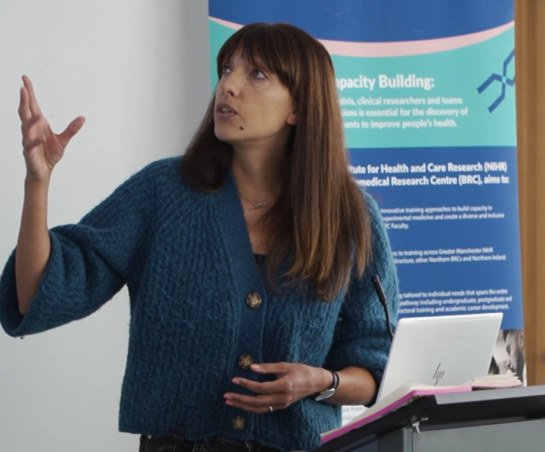 A person giving a talk standing in front of a lectern. They are wearing a teal cardigan and have long brown hair.