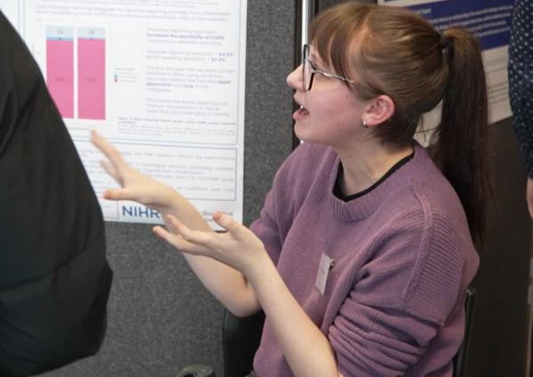 A person discussing their academic poster. They have brown hair worn in a ponytail and are wearing glasses and a purple jumper.